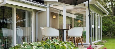 Une maison moderne dotée d’une véranda en verre, de meubles blancs et d’un jardin agrémenté de grandes plantes à feuilles larges.