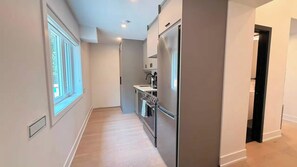 A narrow hallway with a window on the left, a kitchen area with a refrigerator and sink on the right, and a door at the end.