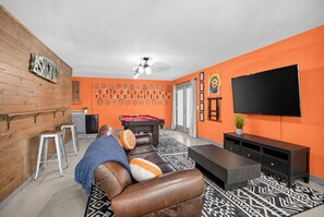 A living room with a pool table, a sofa with orange and white cushions, a television, and a wall with tennis rackets. - Modern Farmhouse w Game Room Near Asheville! (Fairview)