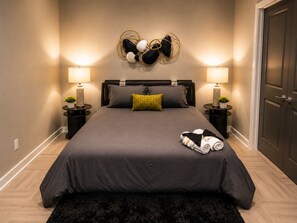 Ein ordentlich bezogenes Bett mit einer dunkelgrauen Tagesdecke, zwei Nachttischlampen und einem dekorativen Wandbild darüber.