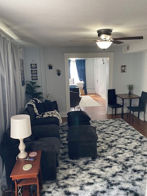 A living room with a sofa, ottoman, lamp, and a black and white furry rug. There is a ceiling fan, a dining area with chairs, and a hallway leading to another room.
