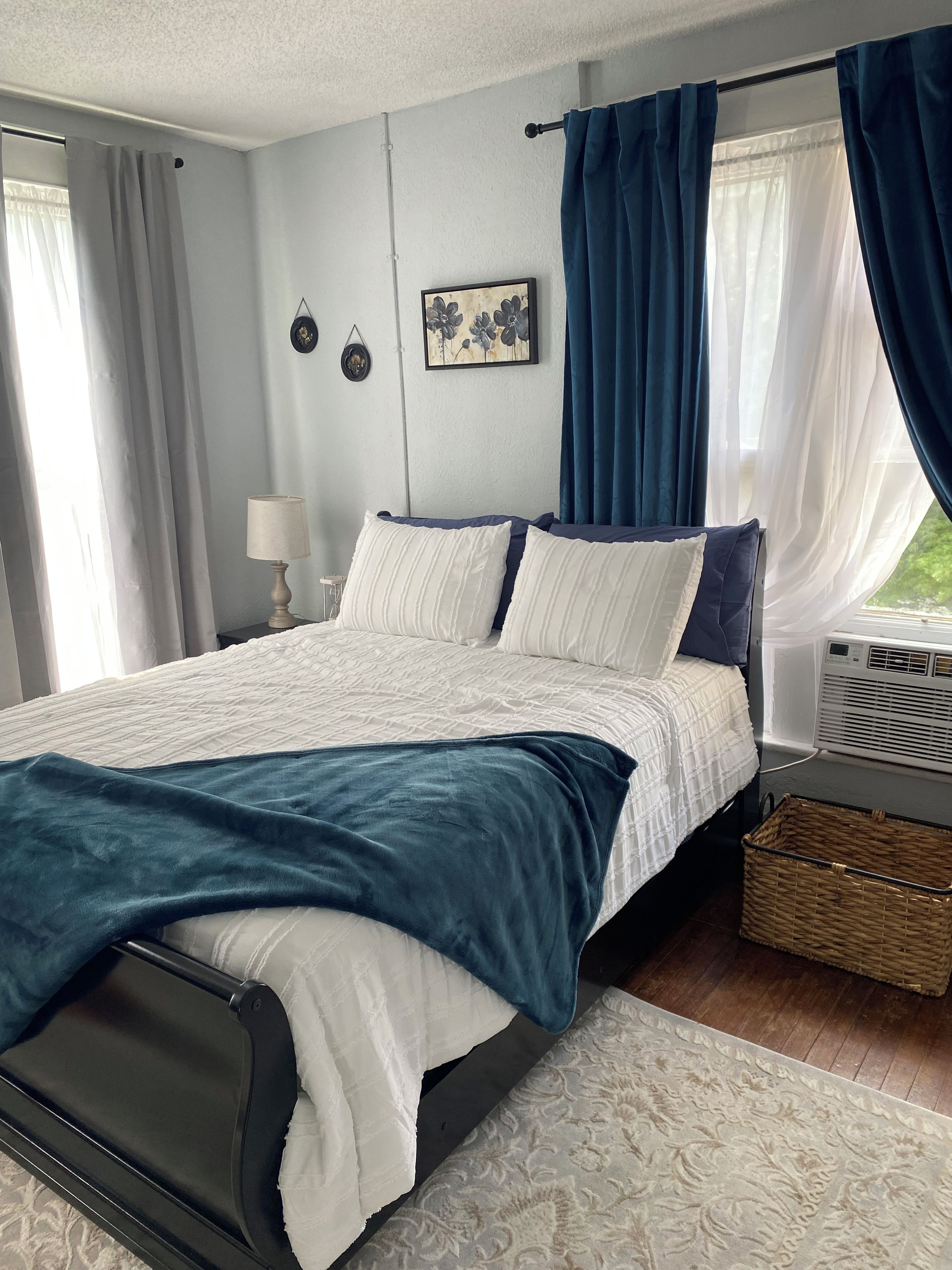 A bedroom with a bed, a bedside table, a lamp, a wall-mounted picture, and a window with curtains.