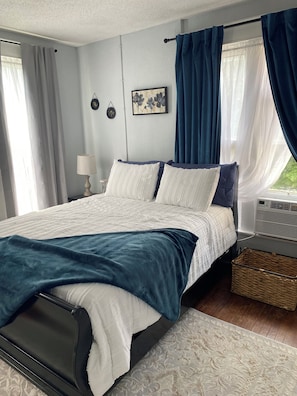 A bedroom with a bed, a bedside table, a lamp, a wall-mounted picture, and a window with curtains.