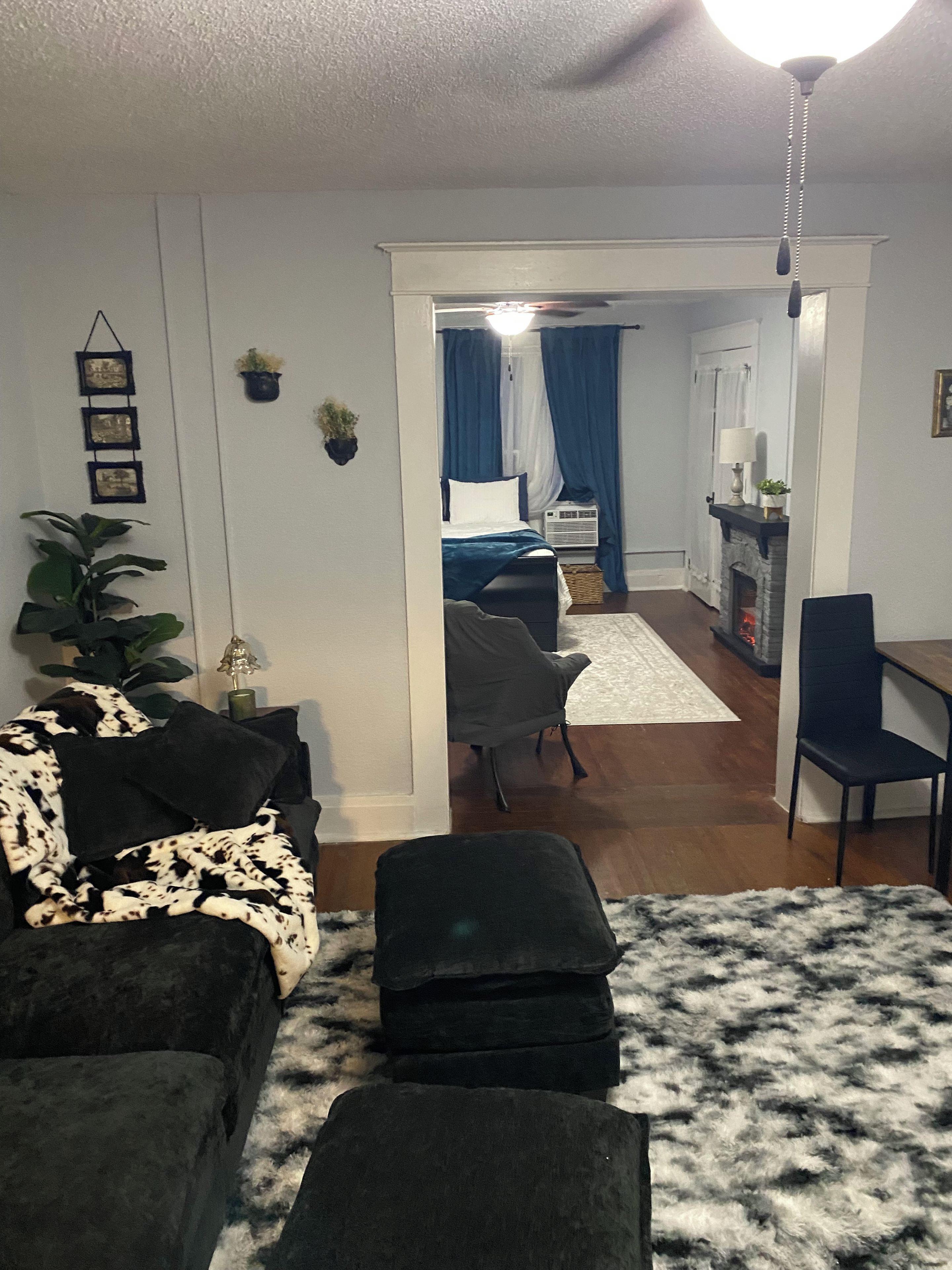 Una sala de estar con un sofá, un reposapiés y un tapete estampado. Se ve un acceso a un dormitorio con una cama y un aparador.