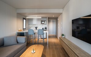Una cocina moderna con horno empotrado, un comedor con mesa de madera y dos sillas, y una sala de estar con sofá y televisor montado en la pared.