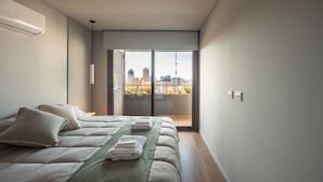 Un dormitorio moderno con cama, almohadas, colcha verde y vista a un paisaje urbano a través de una puerta corrediza.
