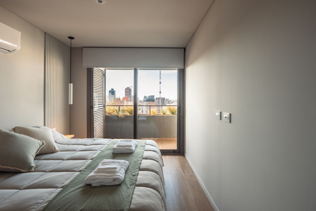 Een moderne slaapkamer met een bed, kussens, een groene sprei en uitzicht op een stadsgezicht door een schuifdeur.