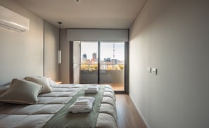 Een moderne slaapkamer met een bed, kussens, een groene sprei en uitzicht op een stadsgezicht door een schuifdeur.