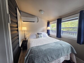 A bedroom with a bed, a nightstand, a lamp, a window with curtains, and an air conditioning unit. - Rustic Riverfront Getaway — Close to Floating, Caverns, Golf & Route 66 (Cuba)