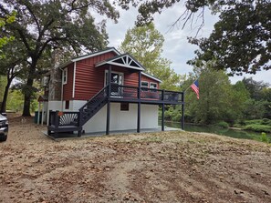 Une cabane à deux étages avec un balcon, entourée d’arbres et bordée d’un étang.