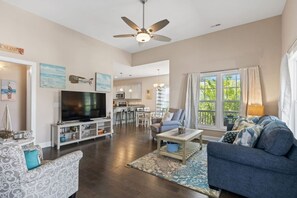 A spacious living room with a flat-screen TV, a ceiling fan, and a large window with curtains. - The Sea Turtle Hideaway𓇼Central+Cozy+Near Beach (Oak Island)