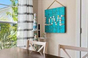 A dining area with a table, chairs, a striped curtain, a lighthouse decoration, and a wall art piece with shells and the word 'LOVE'. - The Sea Turtle Hideaway𓇼Central+Cozy+Near Beach (Oak Island)