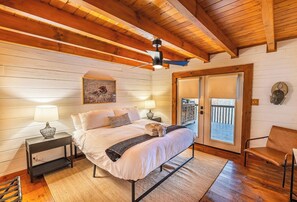Un dormitorio con cama, mesitas de noche, ventilador de techo, ventana y puerta que da a un balcón.
