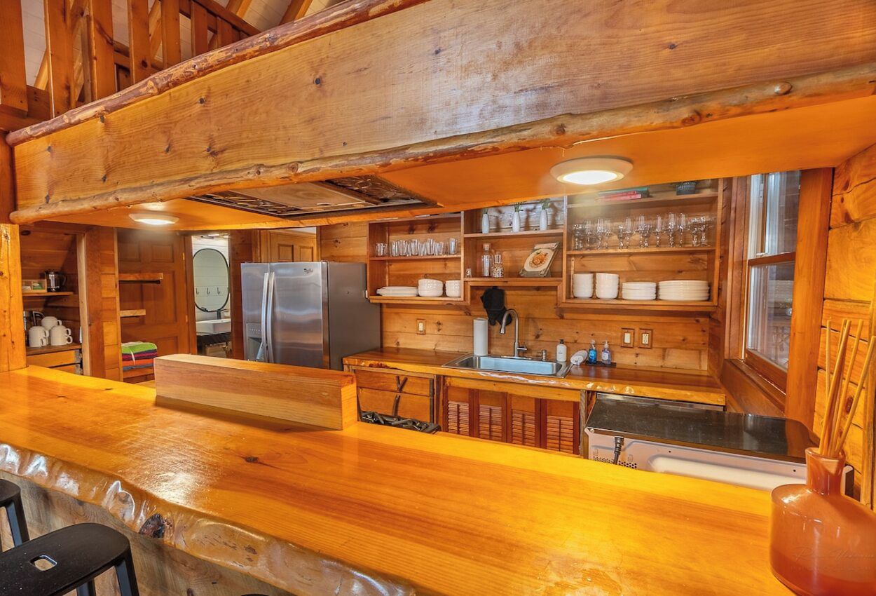 Interno di una baita in legno, con angolo cottura che include un frigorifero, un lavello e mensole in legno con stoviglie e bicchieri.