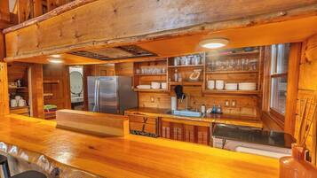 Interno di una baita in legno, con angolo cottura che include un frigorifero, un lavello e mensole in legno con stoviglie e bicchieri.