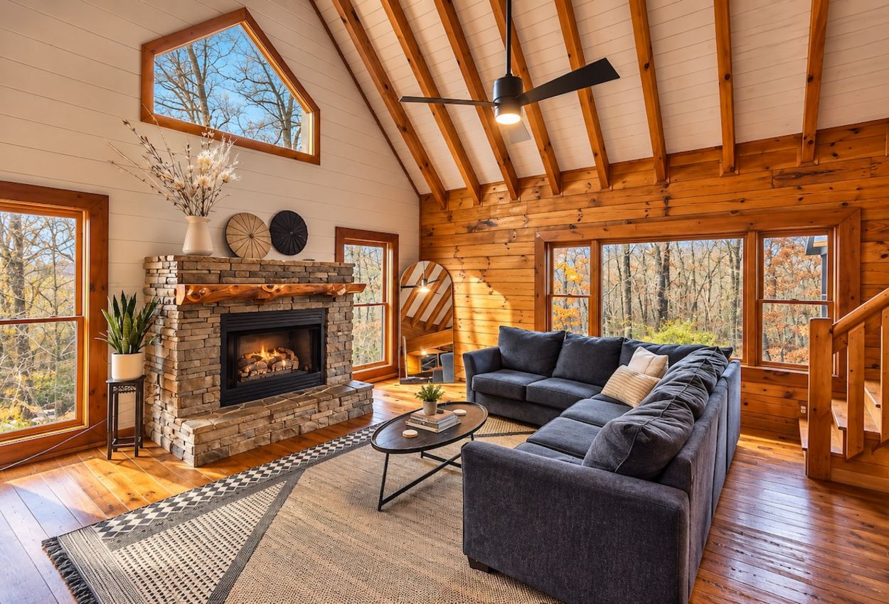 A cozy cabin interior with a stone fireplace, a sectional sofa, and large windows offering a view of the forest.