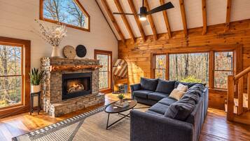 A cozy cabin interior with a stone fireplace, a sectional sofa, and large windows offering a view of the forest.