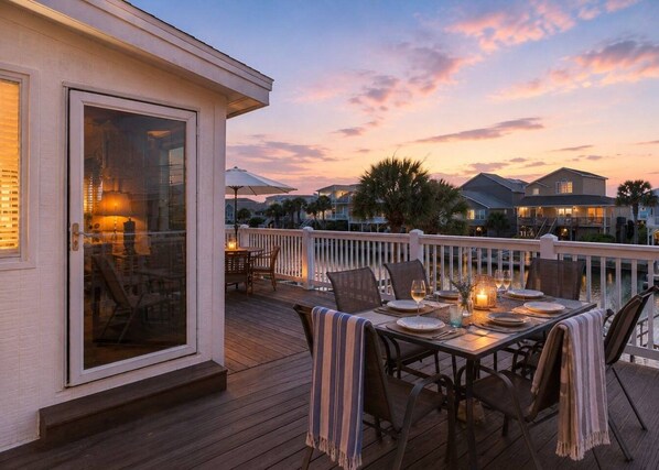 A deck with a table set for dining, overlooking a body of water and a row of houses. - Cakewalk𓇼Canal Front+Private Dock+Walk to Beach (Ocean Isle Beach)