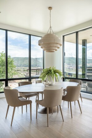 A dining area with a round table, six chairs, a hanging light fixture, and a view of a landscape through large windows. - Deer Valley East Village - Mountain Views - Sleeps 12 4B/5.5B - Ski in Minutes (Daniel)
