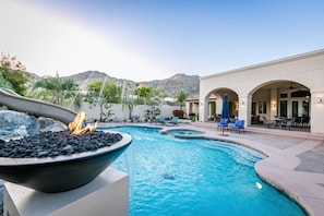 A swimming pool with a fire feature, lounge chairs, and a patio area with tables and umbrellas. - Desert Paradise: Waterslide, Pickleball, Basketball, Game Room, & Gym! (Palm Desert)