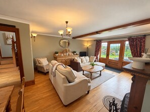 A spacious living room with a wooden staircase, a dining area with a round table, and a large window with curtains. - Ty Cerrig Bach – Hot tub, games room & pet-friendly stay near Benllech (Tyn-y-Gongl)