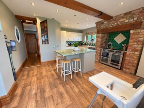 Una cocina con suelo de madera, una isla central con taburetes, pared de ladrillo y salpicadero verde, y un horno moderno.