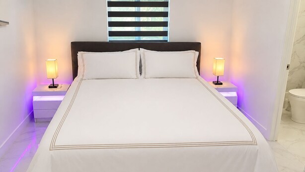 A neatly made bed with a white bedspread and two pillows, flanked by bedside tables with lamps, in a room with a window and a toilet visible. - Private King Suite with Private Entrance & Parking in Hialeah (Hialeah)