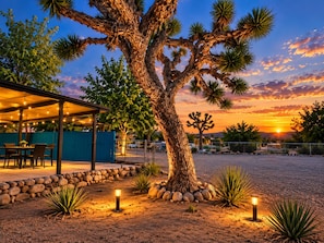 A covered outdoor seating area with tables and chairs, a large tree, and a sunset in the background. - Casa Sol, Fire Pit, Games, BBQ, Stargazing, More (Yucca Valley)
