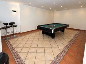 Ein Billardtisch mit grüner Spielbahn, umgeben von einem Teppich mit Musterbordüre, und zwei Barhocker mit schwarzen Sitzen und silbernen Beinen.