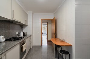 Eine schmale Küche mit Gasherd, Toaster und Spüle. Ein Holztisch mit zwei Stühlen und einem Drahtkorb steht dort. Die Küche verfügt über weiße Schränke und graue Fliesen.