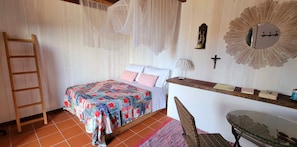 Ein Schlafzimmer mit einem Bett, einer Holzleiter, einem Stuhl, einem Schreibtisch mit Spiegel und einem Kreuz an der Wand.