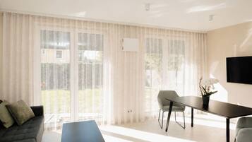 Een moderne woonkamer met een bank, een eettafel, stoelen, grote ramen met dunne gordijnen en een flatscreen televisie aan de muur.