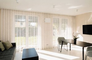 Ein modernes Wohnzimmer mit einer Couch, einem Esstisch, Stühlen, großen Fenstern mit leichten Vorhängen und einem an der Wand befestigten Flachbildfernseher.