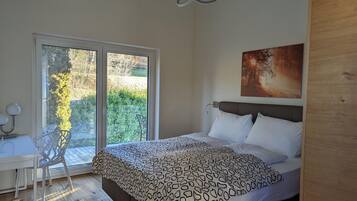 A bedroom with a bed, a desk, and a chair, a large window with a view of greenery, and a painting on the wall.