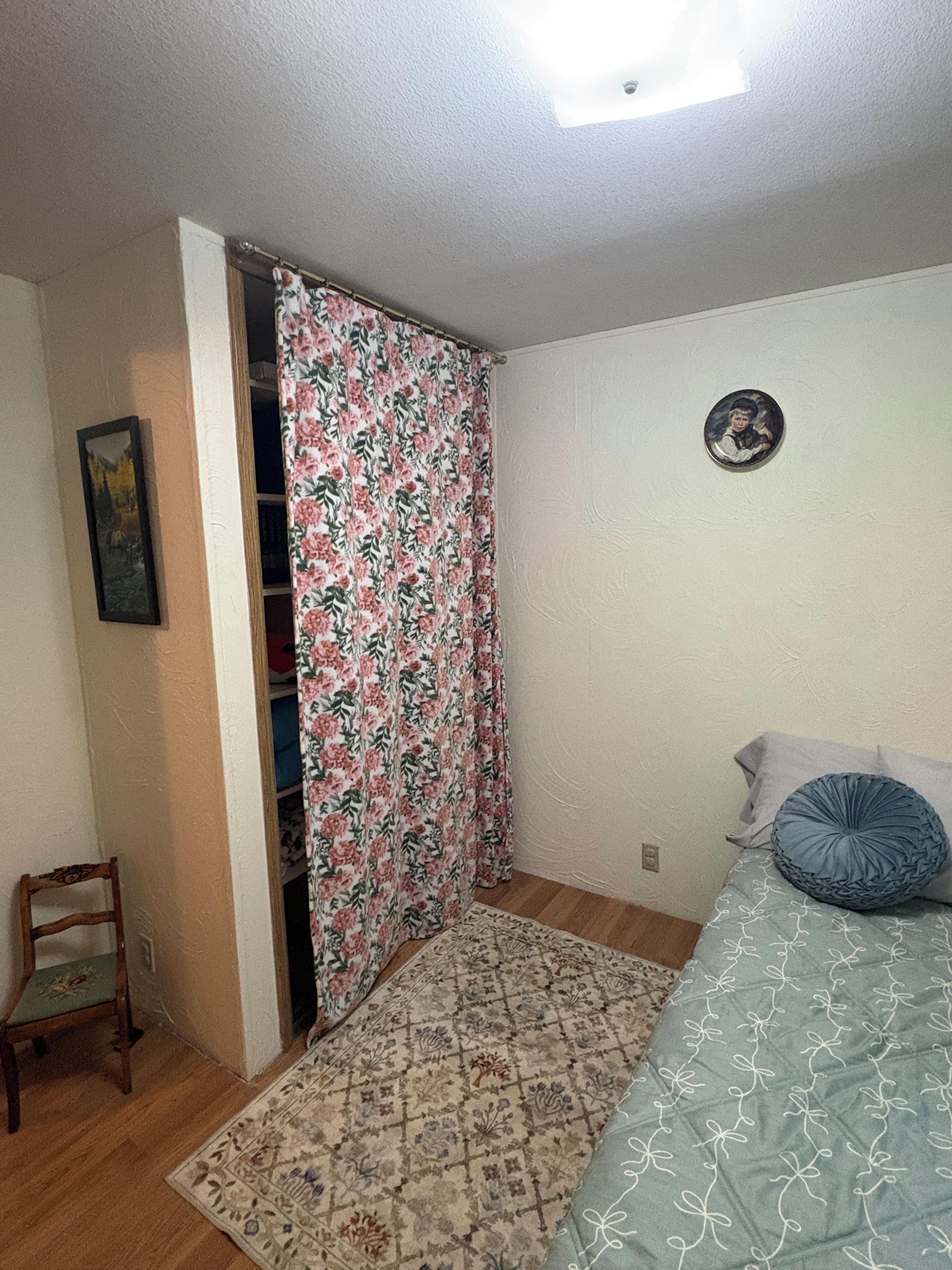 Een slaapkamer met een bed, een stoel, een garderobe en een deur met bloemen gordijnen.