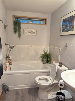 A bathroom with a bathtub, toilet, and sink. There is a window above the bathtub and a plant hanging on the wall. - Peaceful mountain escape nestled in the trees. (Sequim)