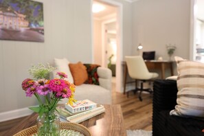 Ein Wohnzimmer mit Couch, Sessel, Schreibtisch, Computern und einer Blumenvase auf dem Couchtisch.