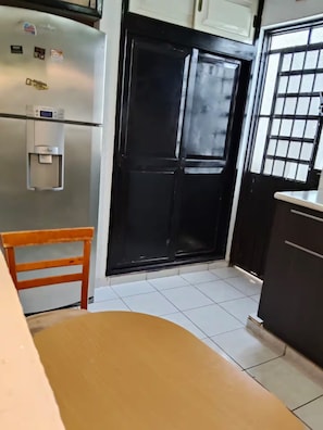 A kitchen with a refrigerator, a water dispenser, a black door, and a tiled floor. - Casa pame! (CHELEM)