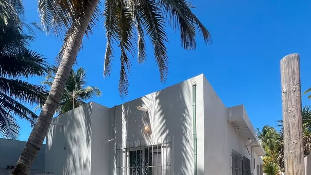 Un bâtiment blanc aux fenêtres grillagées et au sol en béton, entouré de palmiers et surplombé d’un ciel bleu azur.