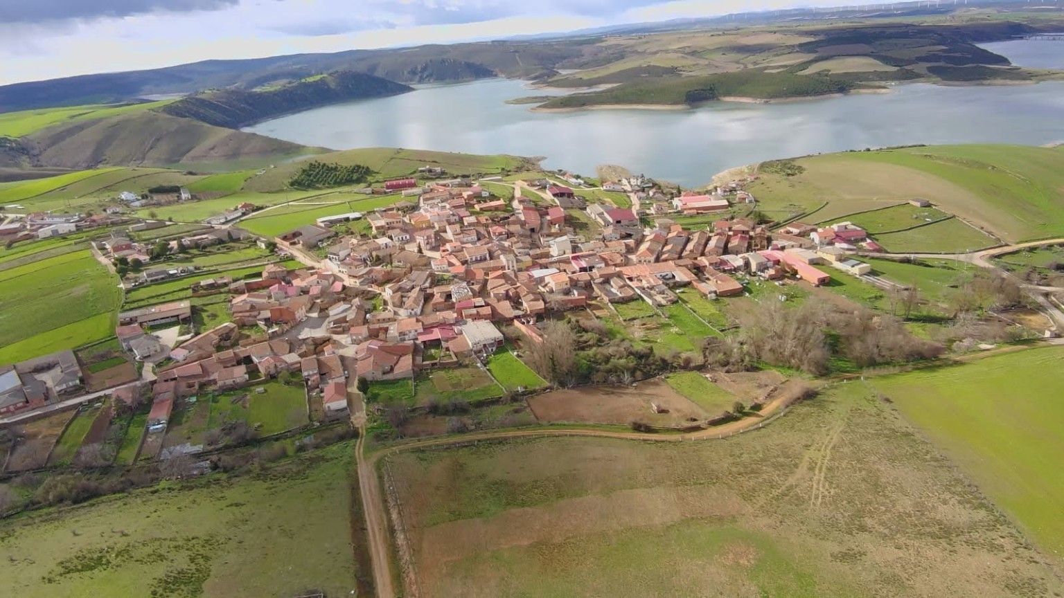 Vista aerea di un piccolo villaggio con case raggruppate attorno a un'area centrale, circondato da campi e un lago.