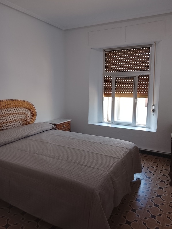 A bedroom with a bed, a nightstand, a wicker chair, a window with a wooden blind, and patterned floor tiles.