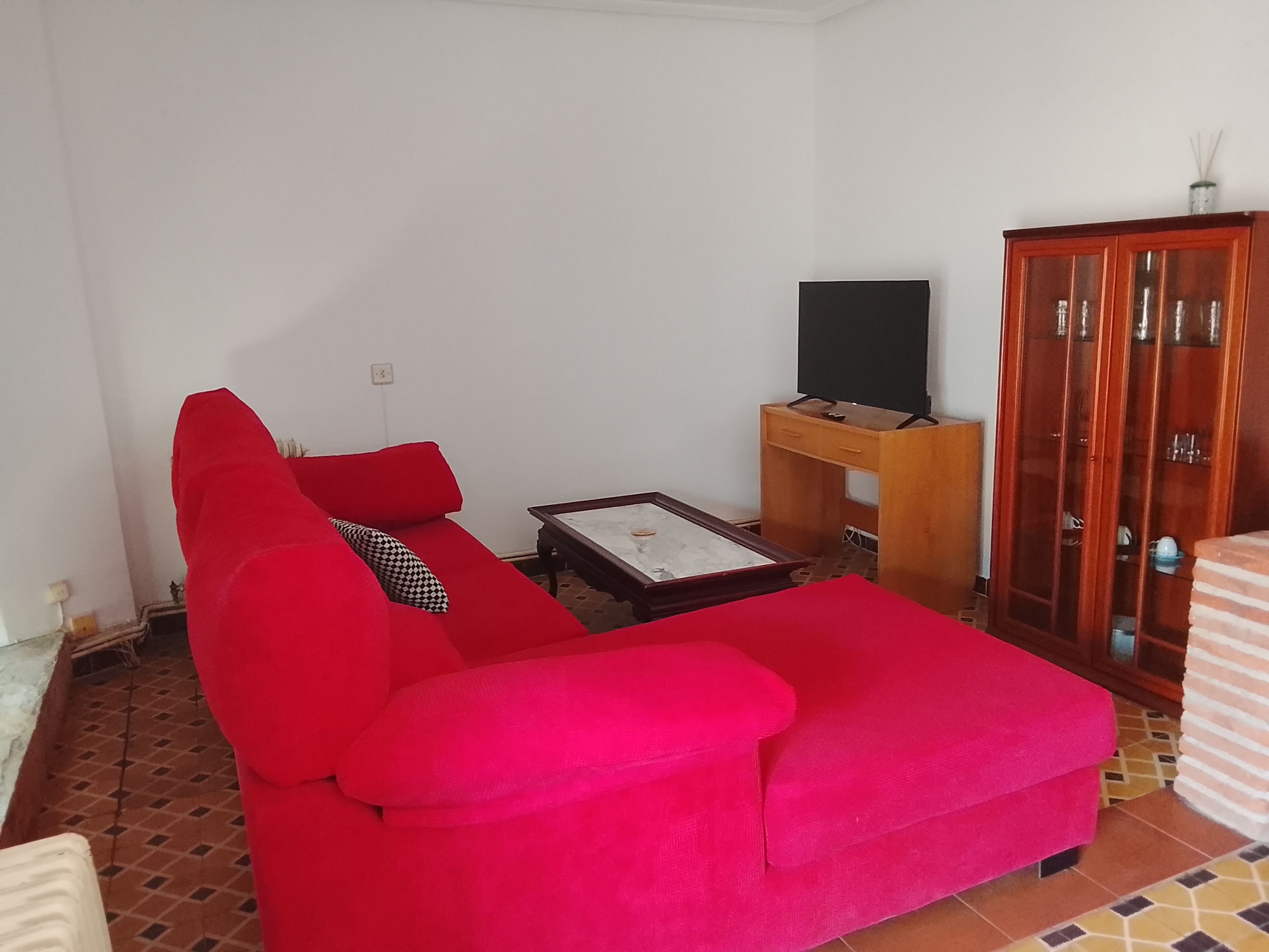 Una sala de estar con un sofá rojo, un mueble de madera con puertas de vidrio y un televisor sobre un soporte.