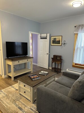 Ein Wohnzimmer mit Fernseher auf einem Schrank, einer Couch, einem Couchtisch mit Büchern, einer Tür und einem an der Wand hängenden Bild.
