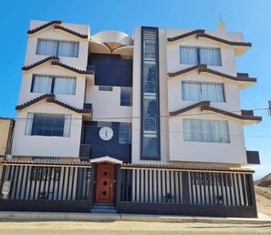Un edificio de varios pisos con un elemento vertical central, balcones y una entrada protegida por una reja.