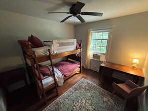 A bunk bed room with a ceiling fan, a desk with a lamp, and a window with curtains.