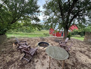 Un espace extérieur aménagé avec des chaises Adirondack et un foyer, entouré d’arbres et avec une grange rouge en arrière-plan.