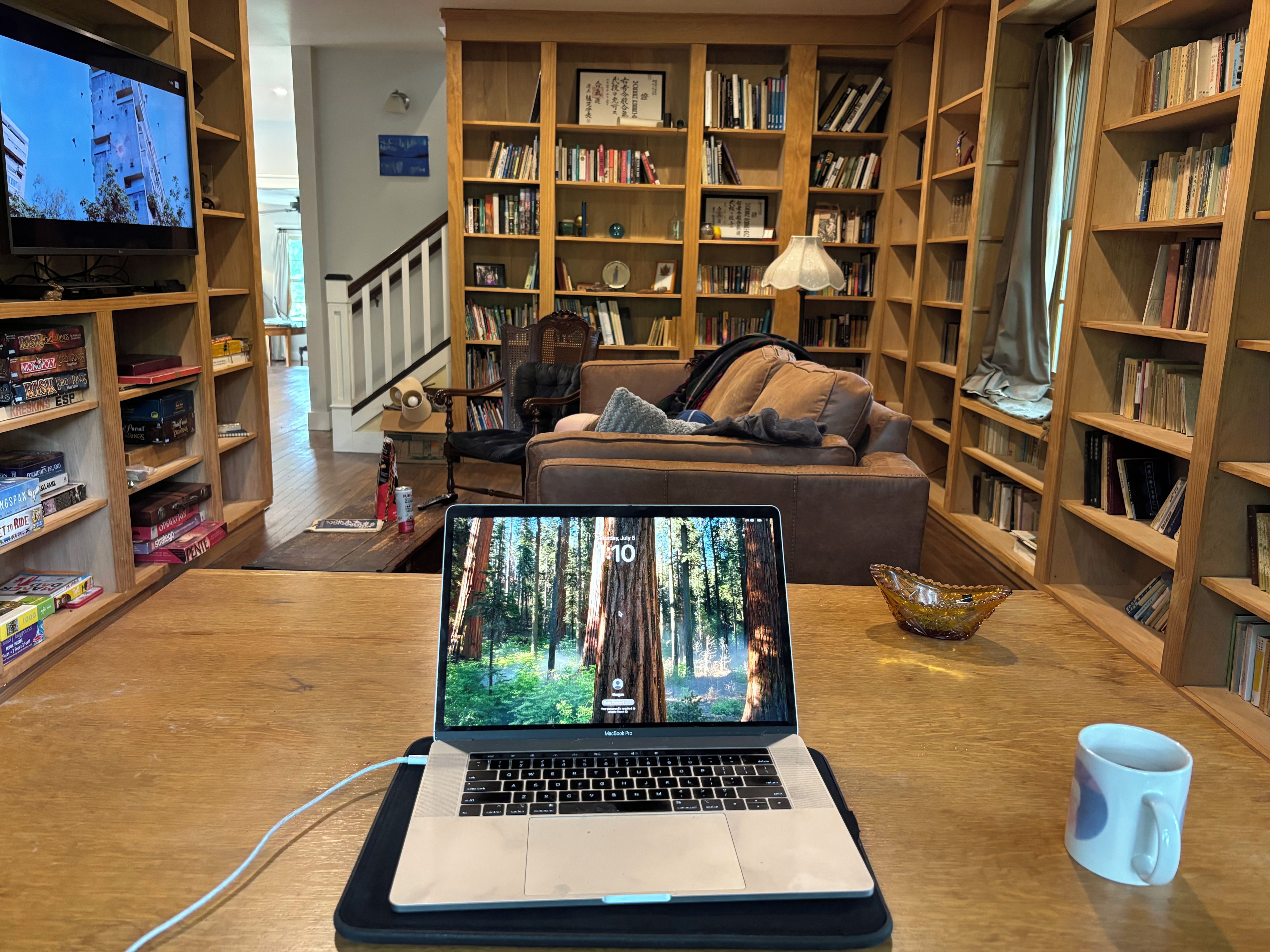 Un salon chaleureux avec un ordinateur portable posé sur une table, entouré d’étagères remplies de livres et d’un téléviseur qui diffuse une image d’un bâtiment.