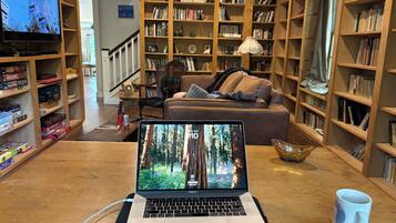 Un salon chaleureux avec un ordinateur portable posé sur une table, entouré d’étagères remplies de livres et d’un téléviseur qui diffuse une image d’un bâtiment.