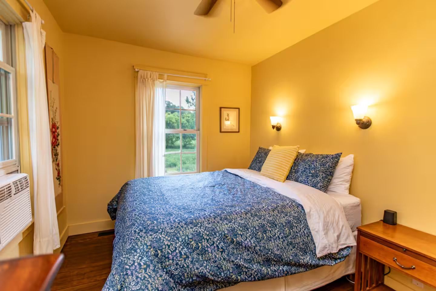 Une chambre à coucher avec un lit, une fenêtre avec des rideaux, une lampe fixée au mur et une table de chevet.