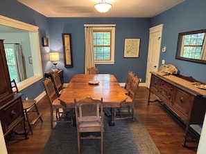 Une salle à manger avec une table en bois, des chaises, un lustre et une vue sur l’extérieur par les fenêtres.